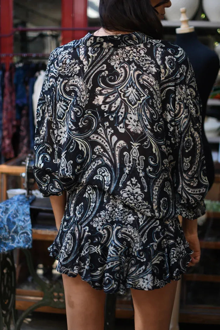 
                  
                    Blusa de gasa con estampado paisley crudo sobre fondo negro y escote en V con cordón, vista de tres cuartos.
                  
                
