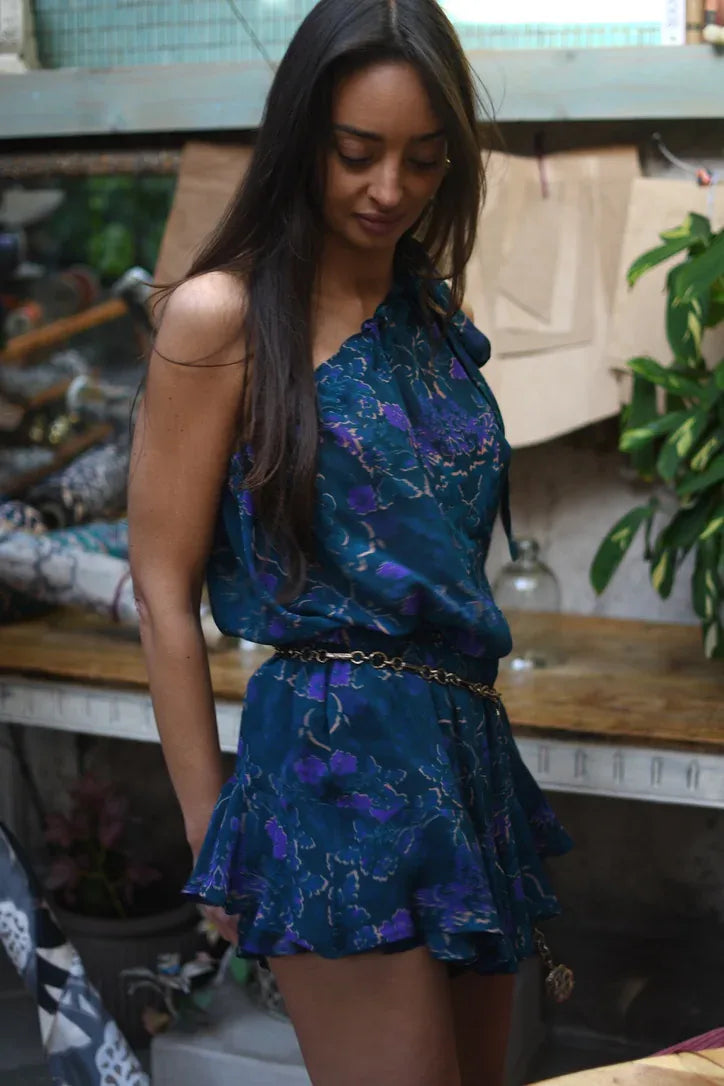 Top de gasa de un solo hombro con estampado floral violeta sobre fondo turquesa, vista posterior.